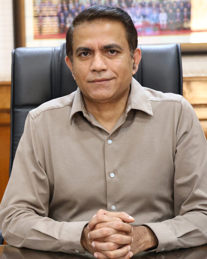 Alok Kumar seated at his desk, hands clasped, in front of a framed group photograph.